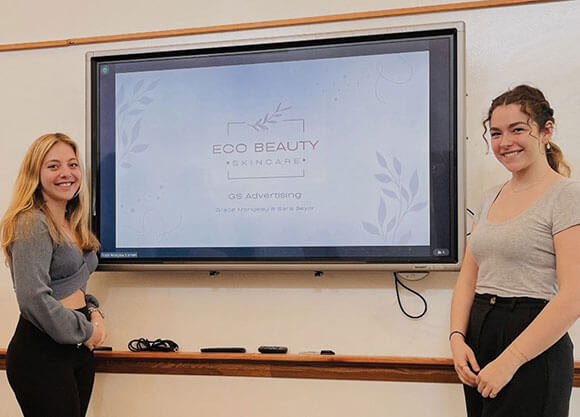 2 students standing next to the title slide of their capstone presentation