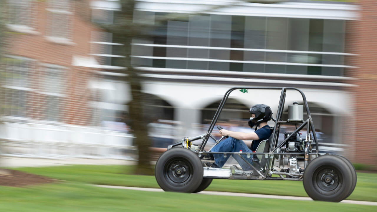 Blurred photo of student driving the Baja vehicle on campus.